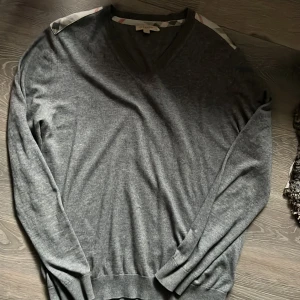 Grå v-ringad tröja från Burberry Brit - Snygg grå v-ringad tröja från Burberry Brit i mjuk cashmere blend. Klassisk passform med långa ärmar och ribbade muddar. Axlarna har Burberrys ikoniska rutiga tygdetaljer som ger en exklusiv touch. Perfekt för dig som gillar stilrena och tidlösa plagg.