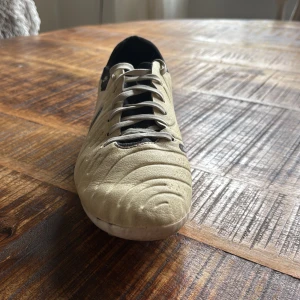 Nike Tiempo Legend 10 fotbollssko, beige - Nike Tiempo Legend 10 fotbollssko i beige med svarta detaljer och snörning. Skon har en rund tå, platt sula med dobbar och är tillverkad i syntetmaterial med FlyTouch-teknologi för extra komfort och kontroll på planen. Perfekt för dig som vill ha stil och funktion på fotbollsplanen.