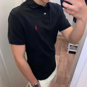 Polo Ralph Lauren (Svart piké) - Säljer en klassisk svart piké t-shirt från Polo Ralph Lauren med röd broderad logga på bröstet. Den är oanvänd och redo for en ny agare. Perfekt for en stilren och sportig look. Pikén är size S men passar även M!