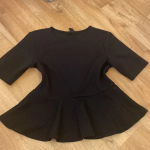 Svart volang topp HM - Säljer en svart volangtop från H&M i storlek M. Toppen har rund halsringning, halvlånga ärmar och markerad midja med utsvängd volang nertill. Materialet är strukturerat och stretchigt, vilket ger en snygg passform. Perfekt till allt!