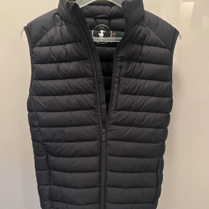 Svart dunväst från Save The Duck - Snygg svart dunväst från Save The Duck i storlek S. Västen är quiltad med horisontella sömmar, har hög krage och dragkedja framtill. Tillverkad i lätt syntetmaterial och är helt animal free. Perfekt för lager-på-lager och stilren look.