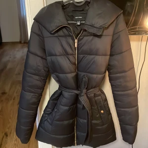 Svart pufferjacka från Vero Moda XS - Svart pufferjacka från Vero Moda i storlek XS. Jackan har bälte i midjan, dragkedja framtill och stora fickor. Snygg, quiltad design med lång ärm och bred krage. Den har tyvärr ett håll på insidan av jackan som inte syns men har endå ett hål! Annars väldigt bra skick!