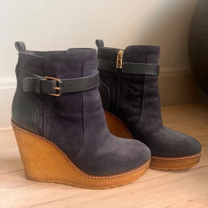 Marinblå wedge boots från Tory Burch - Snygga  wedge boots från Tory Burch i mocka och skinn med guldfärgad dragkedja och spänne. Insidan är fodrad med mjuk päls för extra värme. Sulan är i gummi med tydlig logga undertill och bootsen har en hög kilklack som ger grym höjd. Fint skick inne och ute. 