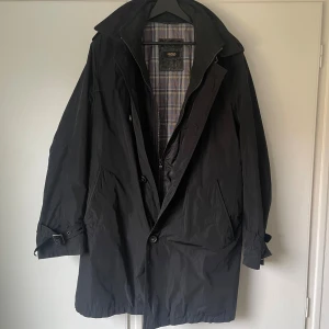 Svart trenchcoat från Ströms Collection | L - Stilren svart trenchcoat från Ströms Collection med klassisk krage, knäppning fram och bälte vid ärmslut. Fodret har rutigt mönster i blått och beige. Jackan har två stora fickor och axelklaffar med knapp. Perfekt för dig som gillar tidlös och clean stil.