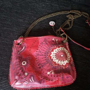 Desigual röd axelväska med kedja - Unik röd axelväska från Desigual med coolt blommönster på framsidan och dekorativa hål. Väskan har en lång kedjerem i antikguld och detaljer i vitt och lila. Tillverkad i syntetmaterial och perfekt för dig som gillar färg och mönster.