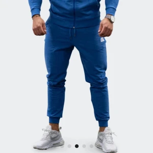 tyngre Blå träningsbyxor - Säljer ett par blåa mjukisbyxor från tyngre med snörning i midjan och resår vid bensluten. Byxorna har en vit logga på framsidan och tryck på ena benet. Perfekta för chill eller träning, med sköna fickor och mjukt bomullsmaterial.