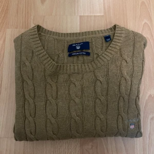 Beige kabelstickad tröja från GANT - Säljer en beige kabelstickad tröja från GANT i premium bomull. Tröjan har rund halsringning, ribbade muddar och GANT-logga broderad på bröstet. Perfekt för dig som gillar klassisk och stilren look.