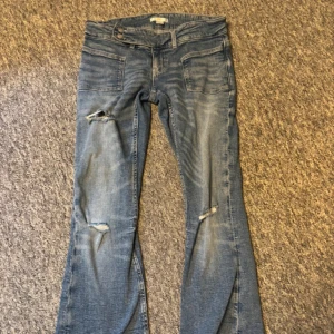 Blå bootcut jeans från Gina Tricot - Bootcut jeans från Gina Tricot i mellanblå tvätt med slitningar och hål på knäna. Klassisk femficksmodell med dragkedja och knapp. Jeansen har en snygg bootcut passform och är tillverkade i jeansmaterial.