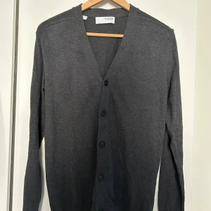 Grå cardigan - Stilren mörkgrå cardigan från Selected Homme med v-ringning och knäppning framtill. Tillverkad i mjuk bomull, perfekt för lager-på-lager. 