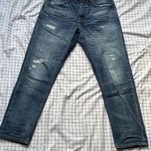 Blå jeans från Nudie Jeans Co - Snygga blå jeans från Nudie Jeans Co med slitna detaljer och klassisk femficksdesign. Jeansen har raka ben, orange kontrastsömmar och knappgylf. Perfekta för dig som gillar en avslappnad och tidlös look. Kostar 1400 nya