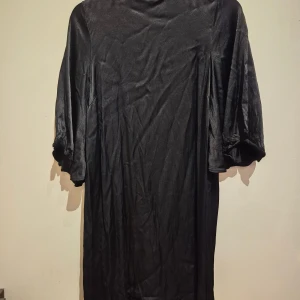 Svart satinklänning Soaked in Luxury - Svart kort satinklänning från Soaked in Luxury med halvlånga ärmar och snygg öppning i ryggen. Klänningen har en lätt glansig finish och en rak, avslappnad passform. Perfekt för dig som gillar stilrena och eleganta plagg.