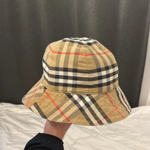 Burberry rutig bucket hat beige - Snygg bucket hat från Burberry i klassiskt rutigt mönster med beige, svart, vitt och röda detaljer. Hatten har mjuk form och är tillverkad i bomull, perfekt för att lyfta din outfit med en ikonisk touch.