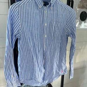 Blåvit randig skjorta från Ralph Lauren - Snygg blå och vit randig skjorta från Ralph Lauren i luftig bomull. Klassisk button-down krage, bröstficka och broderad rosa logga nertill. Långärmad modell med knappar framtill och på manschetterna. Perfekt för en fräsch och stilren look.