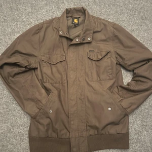 Carthartt overshirt jacka - Snygg mörkgrön jacka från Carhartt med klassisk krage, två bröstfickor med lock och knappar samt ribbade muddar vid ärmslut och midja. Jackan har dragkedja framtill och är tillverkad i slitstark bomull. Perfekt för dig som gillar streetwear nu till hösten