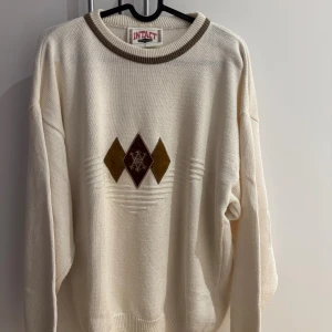 Vit stickad tröja från Intact - Vit stickad tröja från Intact med bruna och beige diamantmönster framtill. Tröjan har ribbade muddar och en kontrasterande brun kant vid halsen. Oversized passform och långärmad, perfekt för lager-på-lager-stil.