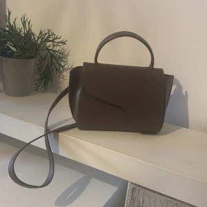 Brun handväska i skinn från ATP Atelier - Snygg höst-brun handväska från ATP Atelier i slätt skinn med stilren design. Väskan har både handtag och avtagbar axelrem med guldfärgade metalldetaljer. Magnetlås framtill och rymligt innerfack. Perfekt för dig som gillar minimalistisk och klassisk stil. Perfekt skick då den är väldigt lite använd!! ☺️🍂🍂