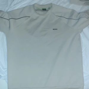 Beige sweatshirt från BOSS - Snygg beige sweatshirt från BOSS med svart logga på bröstet och svarta detaljer längs ärmarna. Tröjan har rund halsringning och långa ärmar. Perfekt för en clean och stilren look. Helt nyt, jätte bra skick och har använt den 5 gånger max.