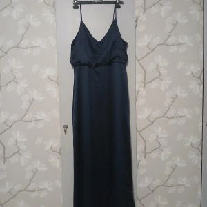 Mörkblå v-ringad långklänning med smala axelband - Elegant mörkblå långklänning med smala axelband och v-ringad hals. Klänningen har en slät, glansig yta och ett stilrent, minimalistiskt snitt som ger en snygg siluett. Perfekt för dig som gillar en klassisk och enkel look.