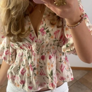 Blommig blus med puffärm och v-ringning - Supersöt blommig blus i vitt med rosa och gröna detaljer. Blusen har djup v-ringning, puffiga trekvartsärmar och smockad midja som ger en snygg siluett. Perfekt för vår och sommar när du vill känna dig extra fräsch.