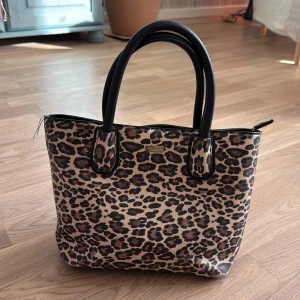 Leopardmönstrad handväska Don Donna - Snygg handväska från Don Donna med leopardmönster i beige och brunt. Väskan har svarta handtag, dragkedja upptill och flera fack inuti samt ett ytterfack med dragkedja. Materialet är syntet med detaljer i svart skinnimitation.