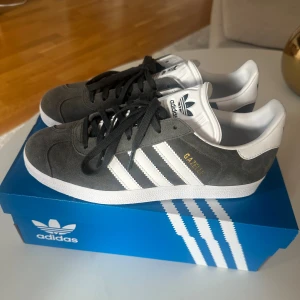 Adidas Gazelle grå sneakers - Snygga Adidas Gazelle sneakers i mörkgrå mocka med vita detaljer och klassiska tre ränder på sidorna. Skorna har svart snörning, vit hälkappa och platt sula. Perfekta för en stilren och sportig look.