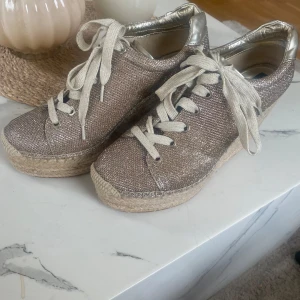 Guldiga platåsneakers från Replay - Säljer ett par glittriga guldiga sneakers från Replay med höga platåklackar i rep och snörning framtill. Skorna har metallic-detaljer vid hälen och en riktigt chunky sula som ger extra höjd. Perfekta för dig som vill sticka ut med din stil.