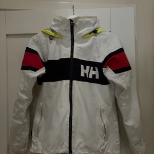 Helly Hansen vit vindjacka med huva - Vit vindjacka från Helly Hansen med svart och röd detalj på ärmarna, stor HH-logga på bröstet och en gul huva. Jackan har dragkedja framtill, två fickor och sportig look. Perfekt för blåsiga dagar och streetstyle.