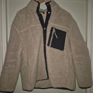 Beige teddyjacka med svart ficka - Mysig beige teddyjacka med hög krage och svart bröstficka med dragkedja. Jackan har en oversized look och är tillverkad i fluffigt fleece-material. Perfekt för kalla dagar när du vill vara både varm och snygg.