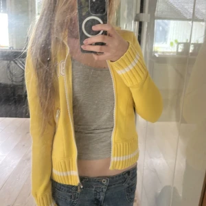 Gul zip up huddie - Gul stickad bomberjacka med vita detaljer och broderade bokstäver och siffror (LS 97) på bröstet. Jackan har ribbade muddar, två fickor med dragkedja. Ger  abercrombie and fitch vibbar .