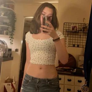 Vit croppad blommig topp - Söt croppad topp i vit färg med små blå blommor. Toppen har korta ärmar, rundad halsringning och en liten rosett framtill. Figurnära passform och stretchigt bomullsmaterial som sitter snyggt på kroppen.