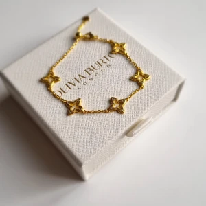 Guldigt armband med blommor - Supersnyggt armband i guldton med fem blommiga länkar dekorerade med små glittriga stenar. Kedjan är tunn och har en smidig karbinlås. Perfekt för dig som gillar detaljer och vill ha något som sticker ut. Lådan i ingår ej.