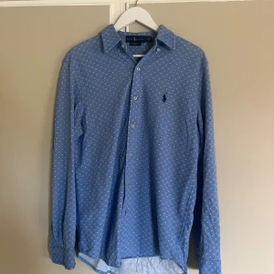 Blå skjorta från Ralph Lauren M - Snygg blå skjorta från Ralph Lauren med små vita och svarta prickar över hela tyget. Klassisk krage, knappar framtill och långärmad modell. Materialet är lätt bomull och har en broderad logga på bröstet. Perfekt för dig som gillar stilrena detaljer.