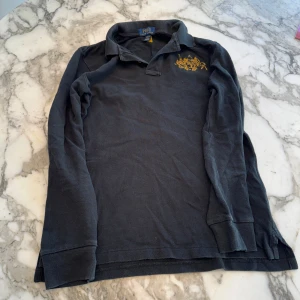 Långärmad piké från Polo Ralph Lauren - Svart långärmad pikétröja från Polo Ralph Lauren med gul broderad logga på bröstet. Storlek 14 - 16 år. Perfekt för dig som gillar stilrena kläder.. 