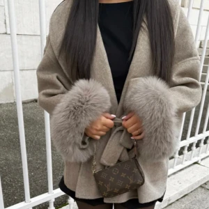 Beige kappa med pälsdetaljer - Snygg beige kappa med pälsdetaljer vid ärmsluten och knytband i midjan. Jackan har en loose passform och ger en lyxig vibe med sina breda ärmar och mjuka material. Perfekt för dig som vill sticka ut med en trendig och elegant look. pälsen är inte äkta , helt ny 