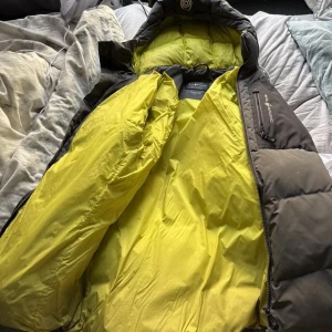 Grå Sail Racing pufferjacka S - Säljer en grå pufferjacka från Sail Racing i storlek S. Jackan har en kontrasterande gul insida, huva och flera dragkedjeförsedda fickor. Perfekt för kalla dagar och har en riktigt schysst streetstil.