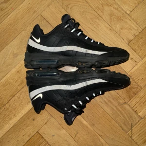 Nike Air Max 95 svarta sneakers - Nike Air Max 95 sneakers i svart med reflekterande silverdetaljer och vit swoosh. Skorna har en chunky sula med synliga luftbubblor och snörning framtill. Ovandelen är i textil och syntet med vågformade paneler. Perfekta för dig som gillar streetwear och sportig stil.