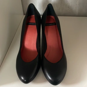 Svarta pumps från Vagabond i skinn - Stilrena svarta pumps från Vagabond i mjukt skinn med rund tå och hög klack. Insidan är fodrad i rött för en snygg kontrast. Perfekta för dig som vill ha en klassisk och tidlös sko med modern känsla.