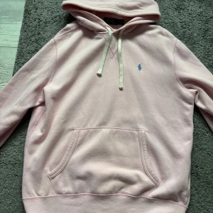 Polo Ralph lauren hoodie  - Ljusrosa hoodie från Polo Ralph Lauren med klassisk blå broderad logga på bröstet. Tröjan har huva med snörning, känguruficka och är tillverkad i mjuk bomull. Perfekt för chill dagar och snygg att styla med jeans eller joggers. Priset är inte hugget i sten så kom med prisförslag.