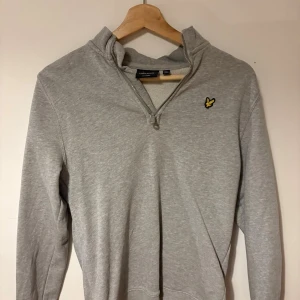 Grå half zip tröja från Lyle & Scott - Grå långärmad tröja från Lyle & Scott med gul logga på bröstet. Tröjan har en half zip-dragkedja vid halsen. Perfekt för en chill och stilren look. Den har används. Men har inte direkt många synliga tecken på användning.