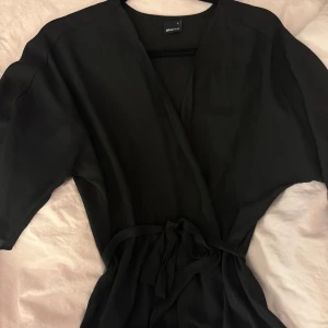 Svart omlottblus från Gina Tricot - Säljer en svart omlottblus från Gina Tricot i storlek S. Blusen har trekvartsärmar, knytband i midjan och en djup v-ringning. Perfekt att styla med jeans eller kjol för en trendig look.