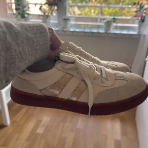 Vita sneakers med gulddeltaljer - Snygga vita sneakers med guld ränder på sidorna och brun sula. Änvands 2 gånger men lite små bara ☺️