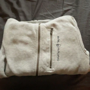 Ljusgrå hoodie från Sail Racing - Säljer en ljusgrå hoodie från Sail Racing med dragkedja och mysig fleecefodrad insida. Tröjan har huva, två dragkedjefickor och broderad logga på bröstet. Perfekt för kyliga dagar och riktigt skön att ha på sig med storlek 150 barn.