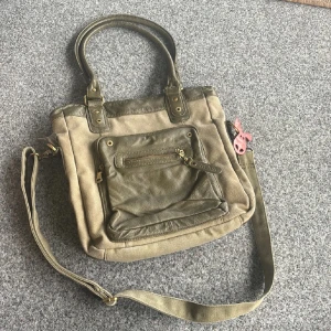 Olivgrön axelväska från NOVA - Cool olivgrön axelväska från NOVA med både handtag och justerbar axelrem. Väskan har detaljer i skinn och canvas, flera dragkedjefack och ett färgglatt blommigt innerfoder. Perfekt för dig som vill ha en praktisk men snygg väska med unik insida. Endast swish betalning skickas spårbart 59kr