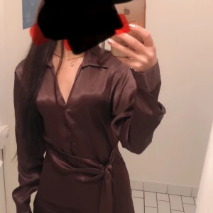 Brun satinklänning med knyt i midjan - Säljer en snygg brun klänning i satin med lång ärm och v-ringning. Klänningen har knappar framtill och ett knytband i midjan som ger en stilren look. Perfekt för dig som gillar en elegant och trendig stil.