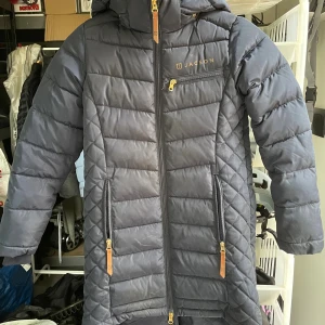 Mörkblå ridkappa från Jacson - Ridkappa från Jacson med snygga läderdetaljer. Jackan har flera fickor med dragkedja och tryckknappar bak. Perfekt för kalla dagar och har en stilren look med diskret logga på bröstet och huvan. Perfekt nu till vintern. Storlek 146/152. Lite lerig men går bort i tvätten!