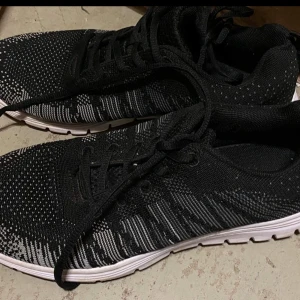 Svarta stickade sneakers strl 40 - Säljer ett par svarta sneakers med vit sula och stickat mönster i svart och vitt. Skorna har snörning och en lätt, flexibel sula med hexagonmönster undertill. Perfekta för dig som gillar sportig och modern stil.