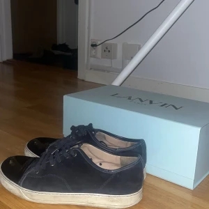 Marinblåa sneakers från Lanvin - Snygga svarta sneakers från Lanvin med klassisk design. Skorna har ovandel i skinn och mocka, rund tå och vita gummisulor. De har svarta snören och stilren look som passar till det mesta. Kommer med originalkartong.