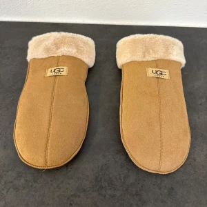 Beige tumvantar från UGC Vogue - Mysiga beige tumvantar från UGC Vogue med mjukt foder och uppvik i fluffig pälsimitation. Utsidan är i mockaliknande material och vantarna har en enkel, stilren design med logga på ovansidan. Perfekta för kalla vinterdagar när du vill hålla händerna varma.