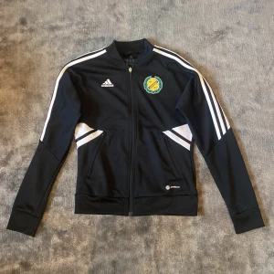 Svart Adidas träningsjacka XS Sävehof - Svart träningsjacka från Adidas med Sävehof märke på bröstet och vita ränder längs ärmarna. Jackan har dragkedja framtill, Stadium-tryck på ryggen och är tillverkad i polyester. Perfekt för dig som gillar sportig stil och handboll 