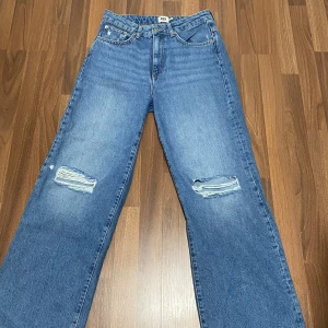 Blåa vida jeans med hål i knäna - Säljer ett par blåa jeans med vida ben och slitna detaljer vid båda knäna. Aldrig använt men har inte lappen kvar. Nypris 300kr storlek M